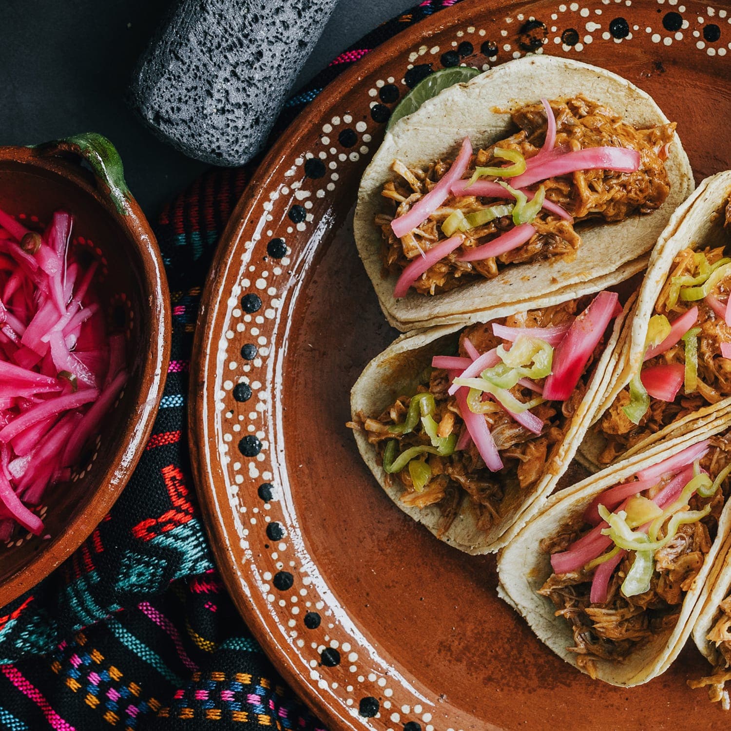 Tacos on a brown plate, sitting on a colorful embroidered tablecloth.